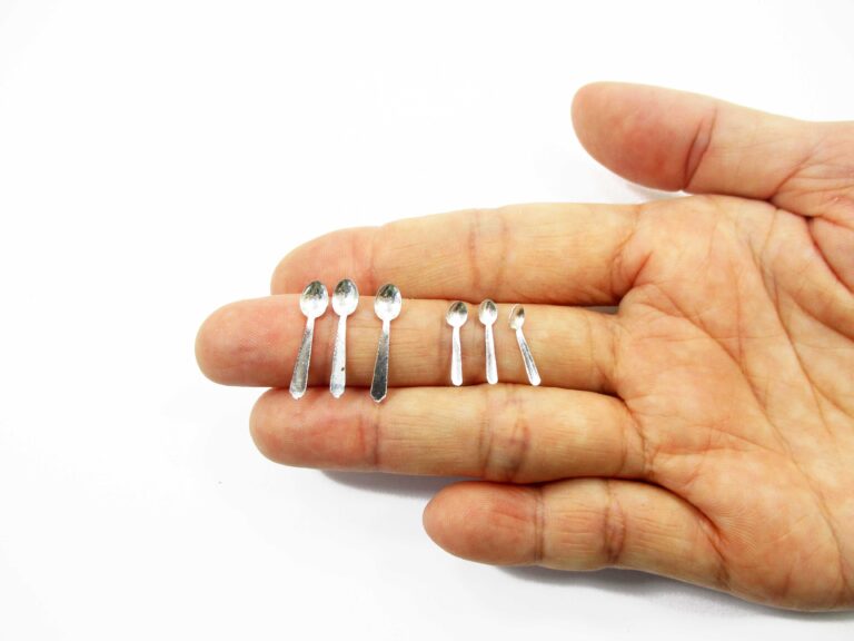 Three 20mm and three 15mm silver-tone tin spoons resting on a person's fingers to show 1:12 scale for dollhouse kitchen decor.